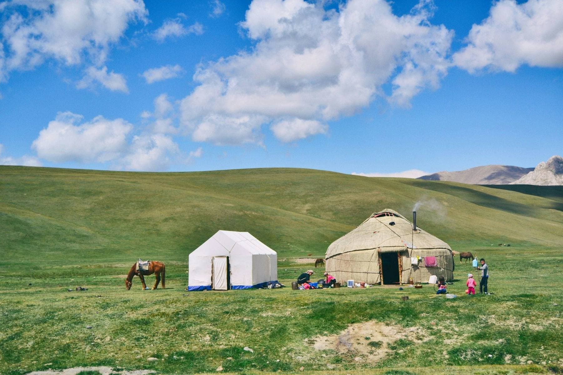 Yurts on the green shore of Song-Köl lake at dawn, with horses grazing and the snow-streaked Tien Shan ridgeline behind