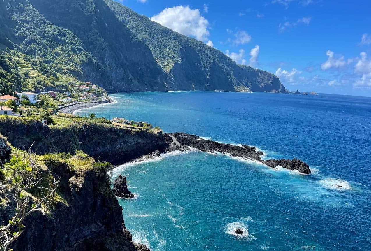 A narrow levada irrigation-channel path cut into a green Madeira mountainside above the clouds, with a hiker on the ridge between Pico do Areeiro and Pico Ruivo