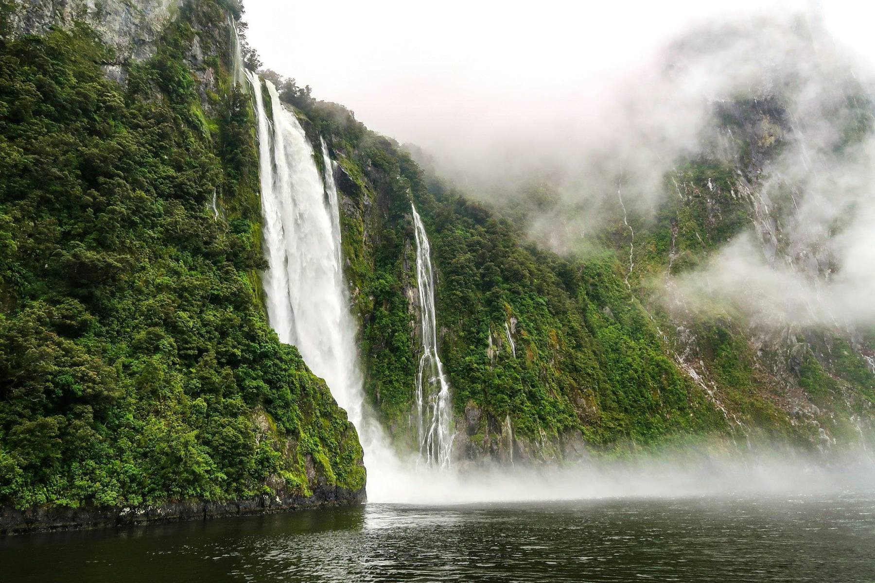 A hiker on a boardwalk in the Clinton Valley on the Milford Track, with waterfalls dropping off sheer Fiordland cliffs and beech forest on either side
