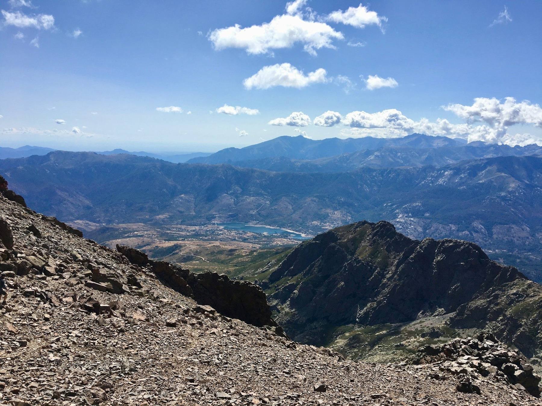 A hiker crossing an exposed granite ridge on the GR20 in Corsica, with jagged peaks and a mountain refuge in the valley below