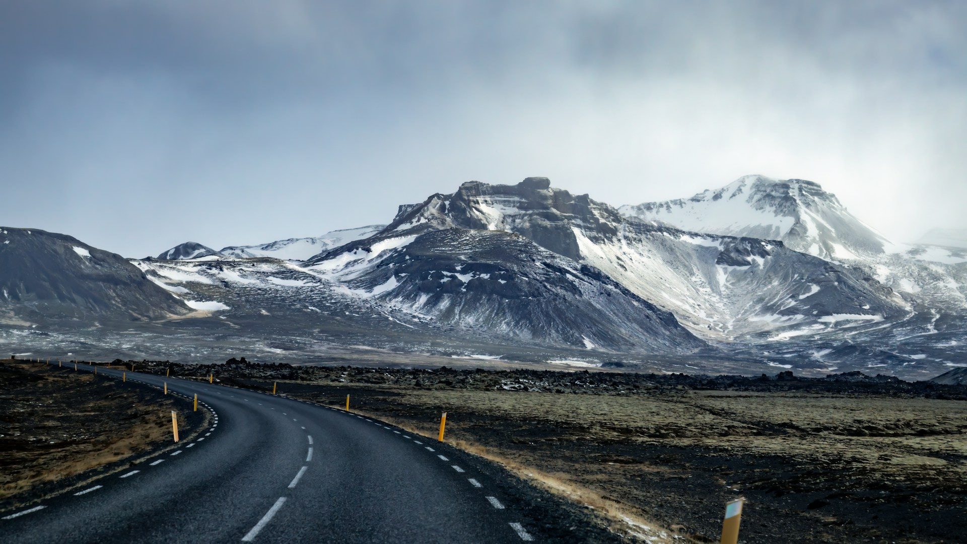 A car on Iceland's Ring Road curving along a black-sand coast beneath a waterfall and low cloud, with a single-lane bridge ahead