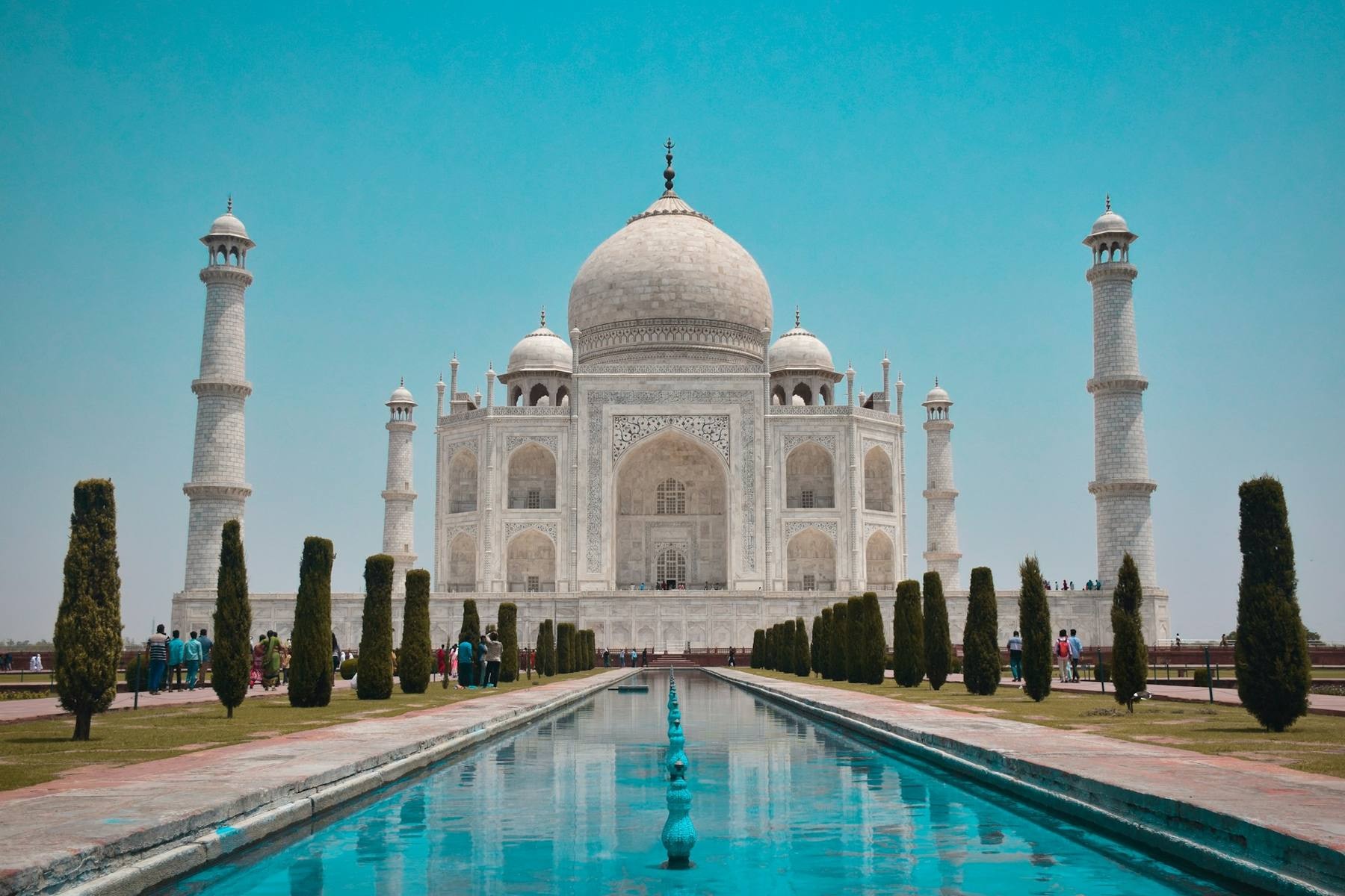 The Taj Mahal at sunrise viewed from across the reflecting pool, with a single traveler in silhouette in the foreground and the broader Golden Triangle circuit implied by a day pack at their feet