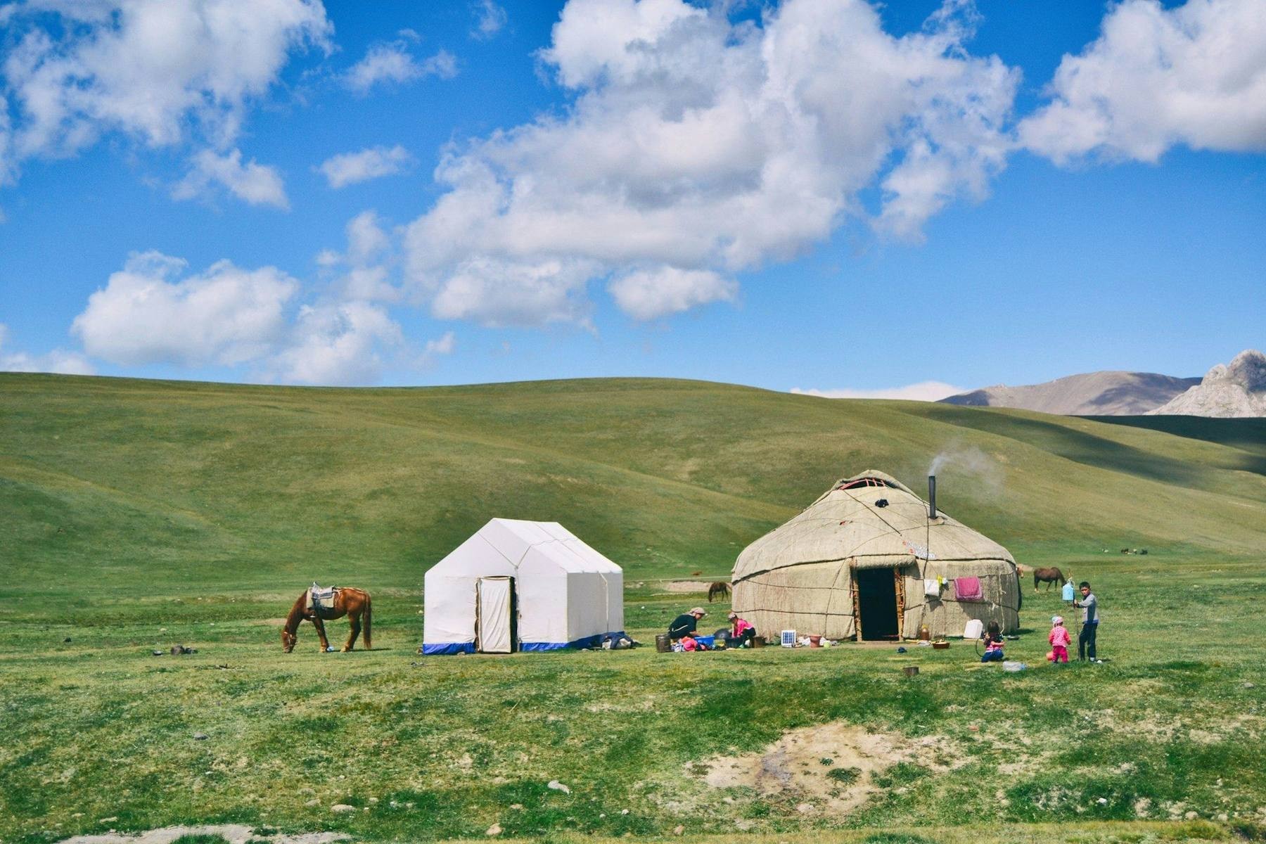 Yurts on the green shore of Song-Köl lake at dawn, with horses grazing and the snow-streaked Tien Shan ridgeline behind