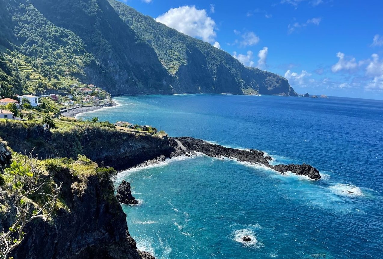A narrow levada irrigation-channel path cut into a green Madeira mountainside above the clouds, with a hiker on the ridge between Pico do Areeiro and Pico Ruivo