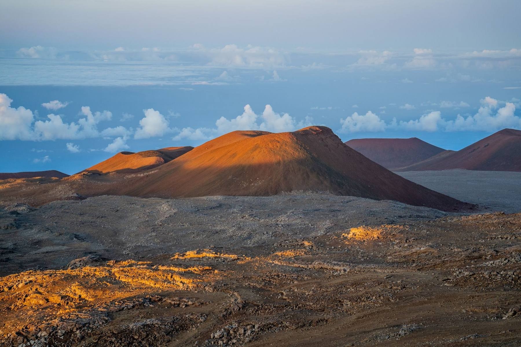 The cinder-cone summit of Mauna Kea above the clouds at sunset, with observatory domes silhouetted against a deep orange sky