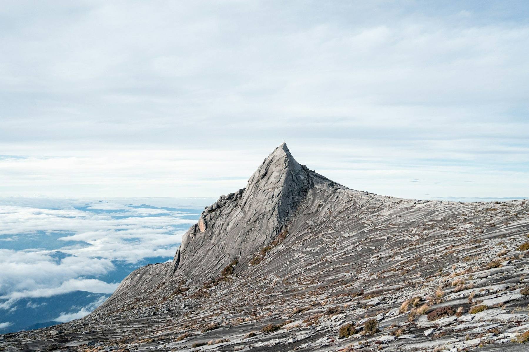 The granite summit plateau of Mount Kinabalu at dawn with climbers crossing the bare rock toward Low's Peak above a sea of cloud, rainforest below
