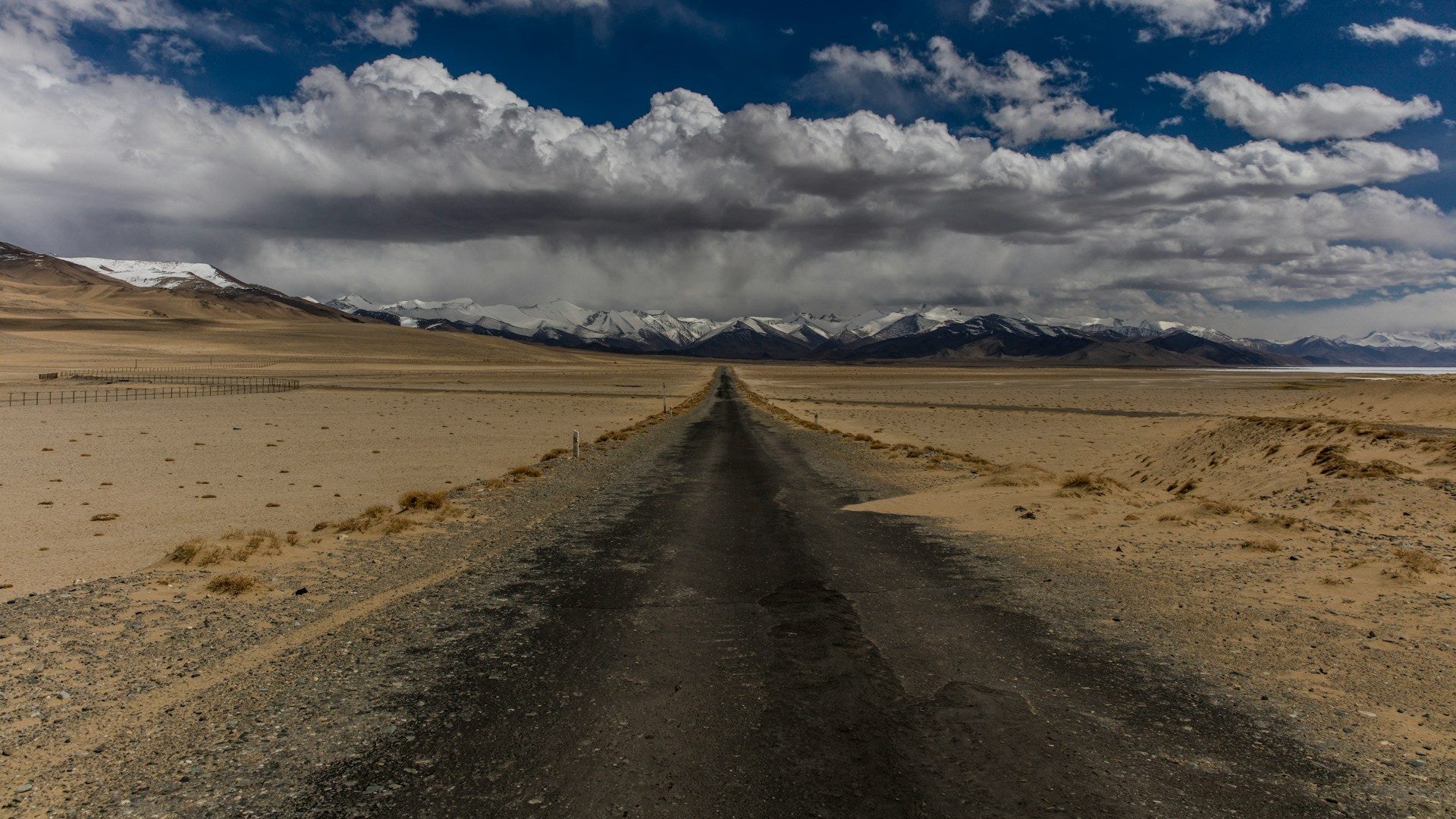 The M41 Pamir Highway crossing the empty high plateau toward Ak-Baital pass, snow-streaked Pamir peaks on the horizon under a hard blue sky