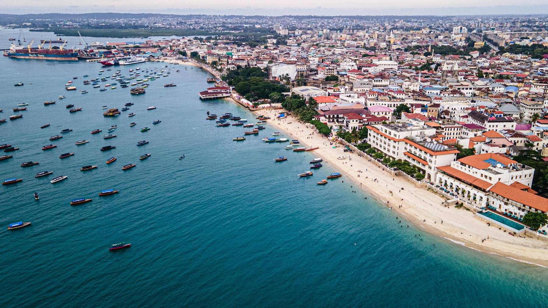 Dhow sailing boats on the turquoise water off Stone Town at golden hour with the old waterfront and palms behind, and a separate inset of a narrow Stone Town alley with carved wooden doors
